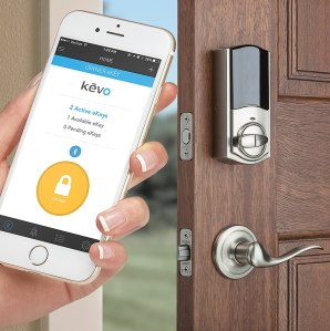 Front door with Kwikset Smart Lock in the background. A hand holding a smart phone showing the Kevo app in the foreground.